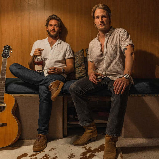 Jonathan Clay and Zach Chance of Jamestown Revival sitting against a wooden wall with cow skin rug at their feet, both holding a whiskey glass. The man on the left is holding a bottle of Oak & Eden’s barrel proof rye whiskey called Round Prairie Rye.