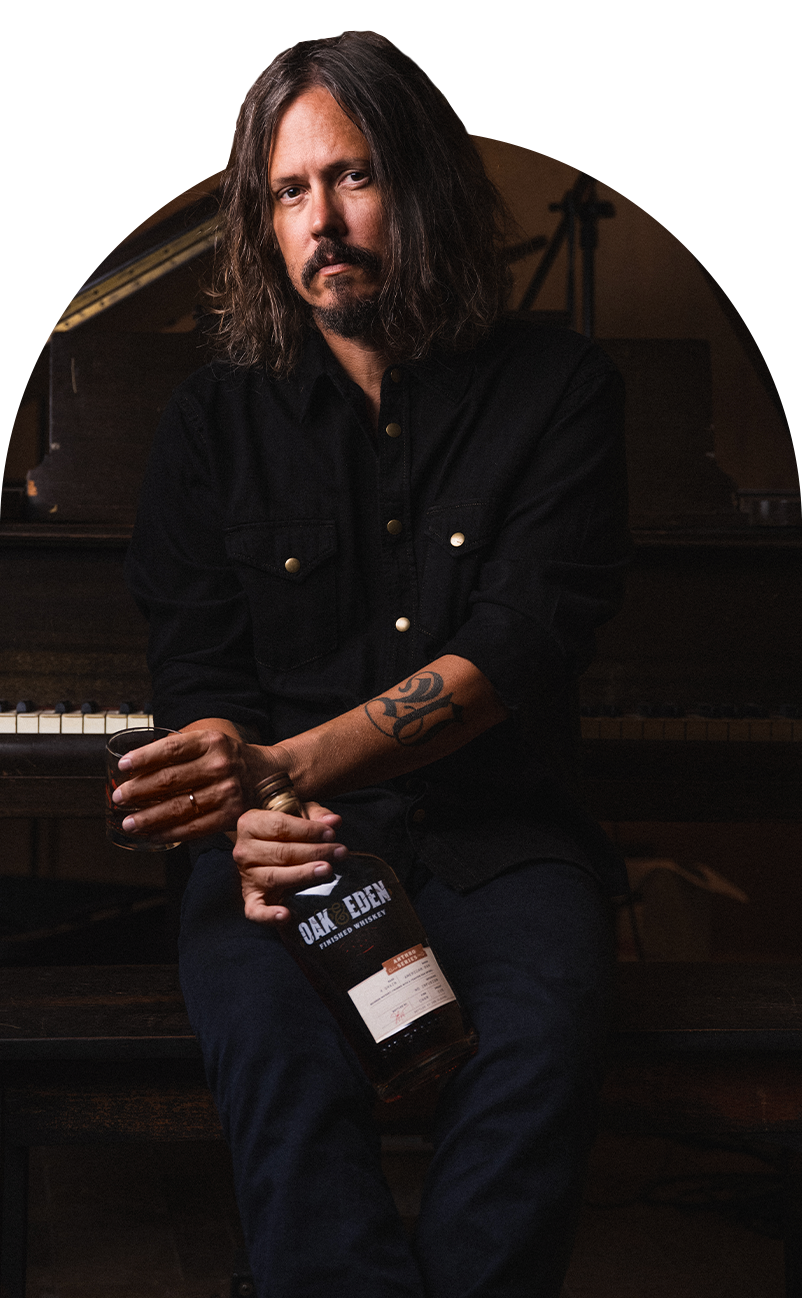 John Paul White from The Civil Wars duet is sitting on a piano bench, holding both a whiskey glass and his custom bottle of Oak & Eden, a barrel strength 4 Grain Bourbon, finished with a charred American Oak spire.