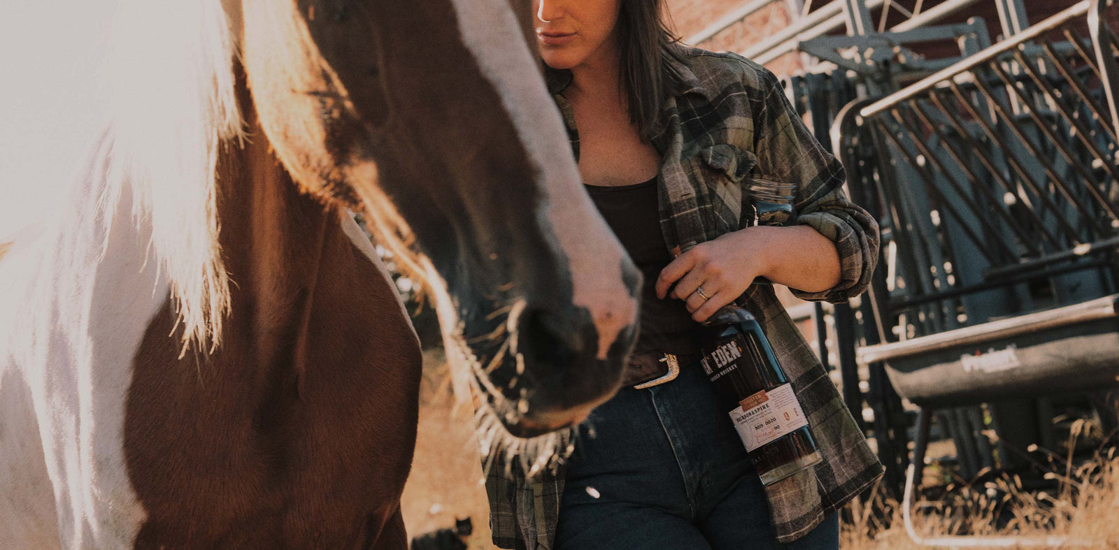 A women standing next to a brown and white haired horse. She is gesturing toward the horse as if she is speaking to it. She is touching the horse with one hand and holding a bottle of Oak & Eden Whiskey in the other hand.
