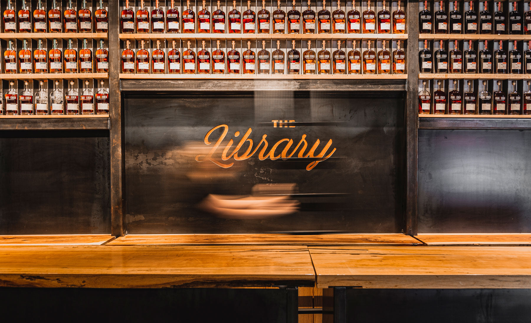 A wooden wall filled with bottles of Oak & Eden whiskey. The text on the wooden wall says The Library in script.
