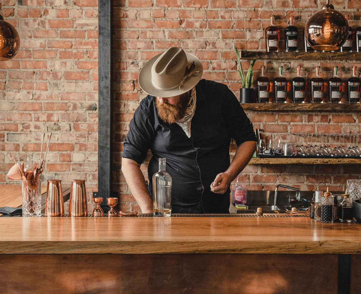 A bartender is standing behind a wooden bar with a brick wall behind him, with multiple shelves filled with Oak & Eden whiskey bottles. The mean is wearing a green hat, and a black shirt with a bandana around his neck, looking down at the bar.
