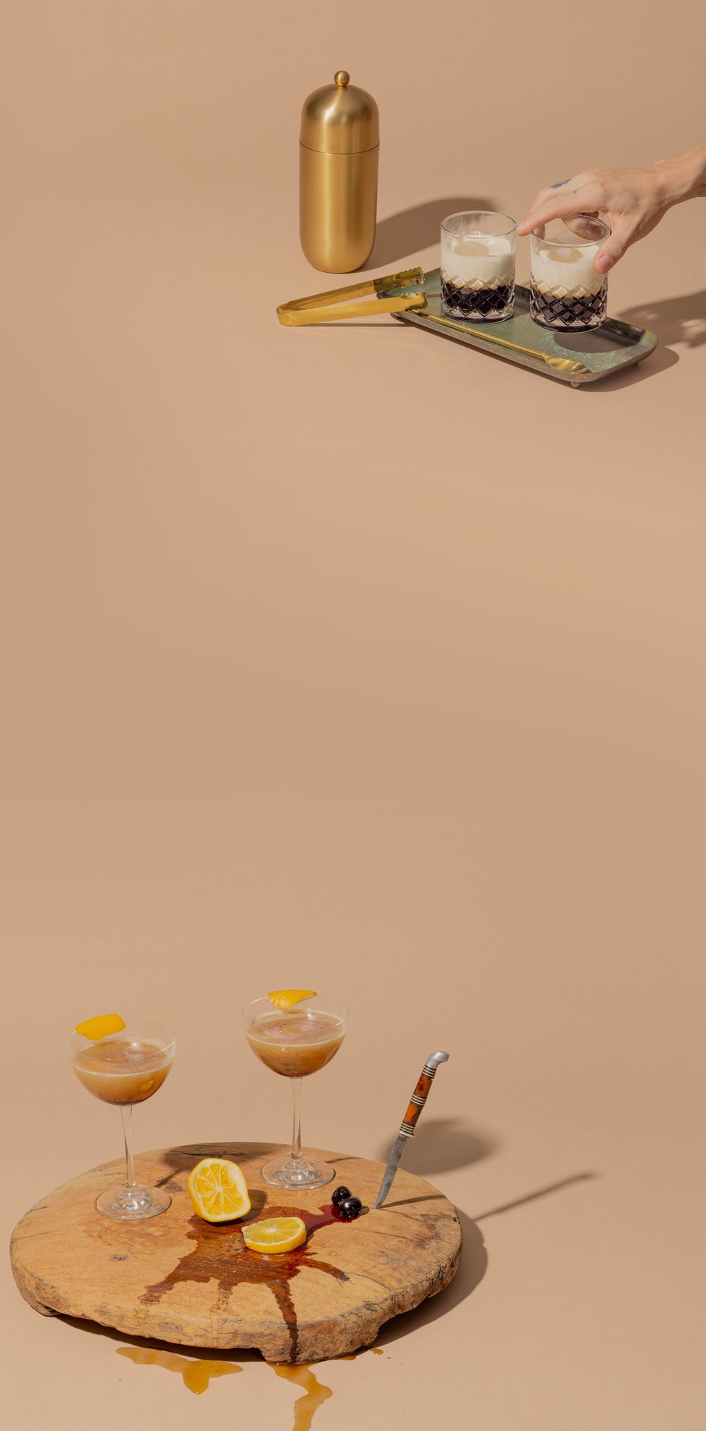 Two cocktail glasses on a wooden cutting board with two orange slices that have juice spilling out underneath, and a knife sticking out of the board. A man setting a cocktail down on a tray with a shaker glass and ice cube tongs next to the tray.