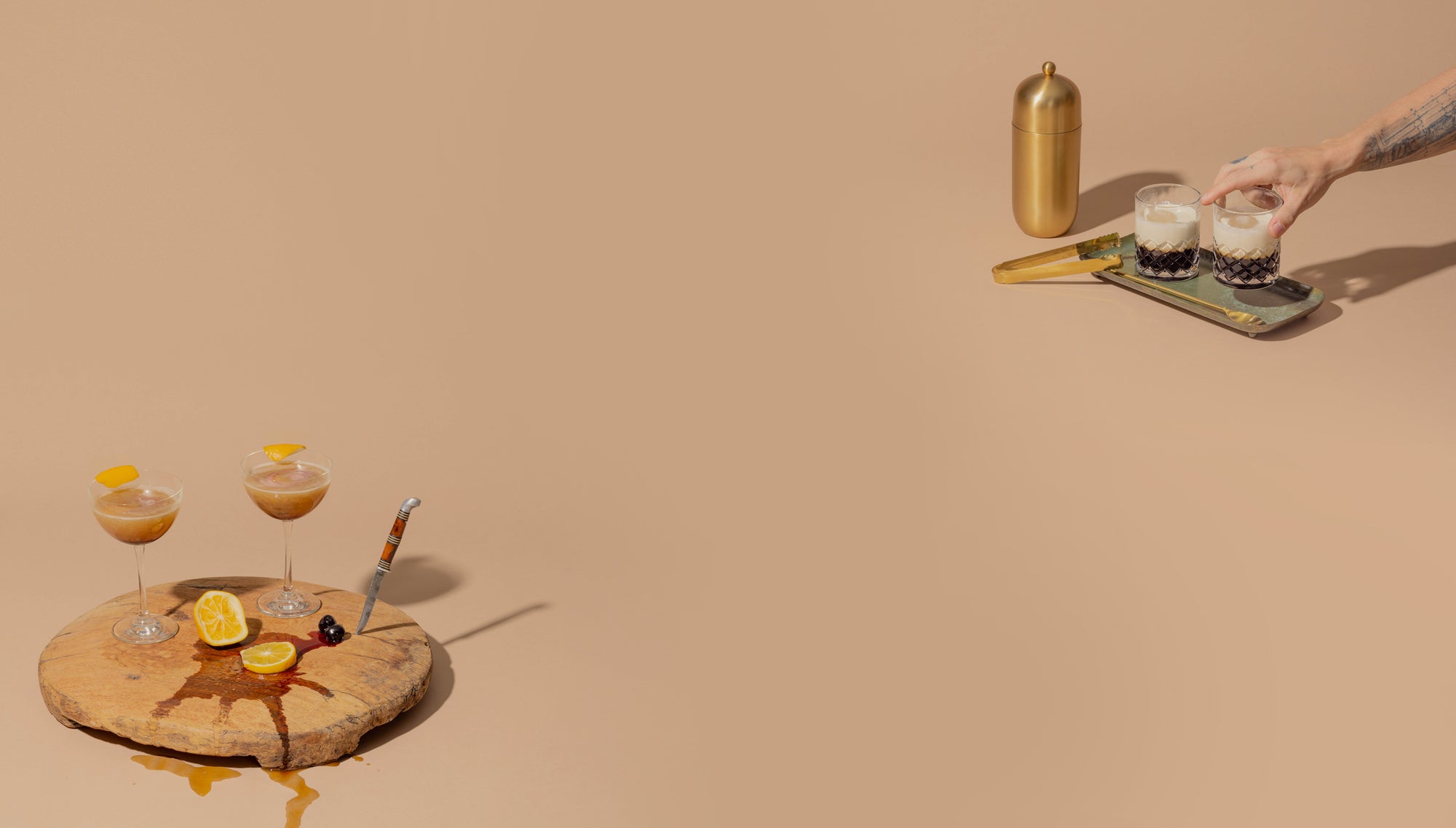 Two cocktail glasses on a wooden cutting board with two orange slices that have juice spilling out underneath, and a knife sticking out of the board. A man setting a cocktail down on a tray with a shaker glass and ice cube tongs next to the tray.