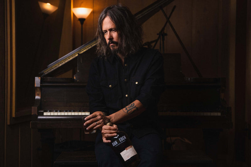 John Paul White from The Civil Wars Americana duet sitting at a piano with a bottle of Oak & Eden Whiskey