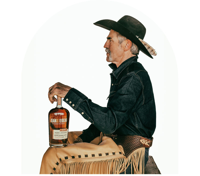 White background featuring Forrie J Smith sitting on a wooden bench holding a bottle of Cold Brew Coffee Infused bourbon that was created in partnership with Oak & Eden Finished Whiskey.