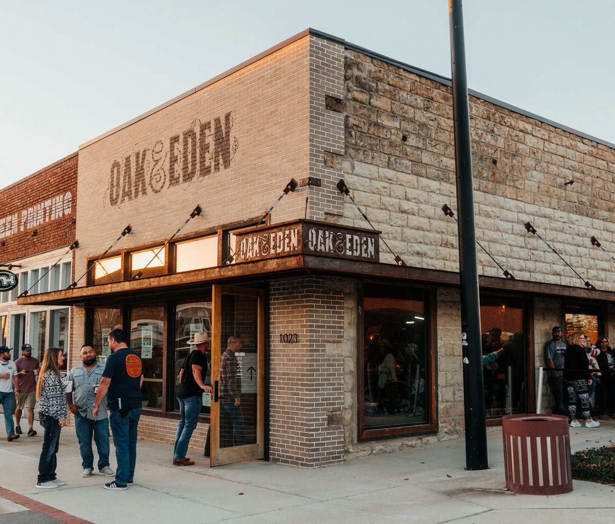 A corner view of a brick building with people walking into it at sunset. The Oak & Eden flagship store sign is visible from the corner of the building and from the front of the building.