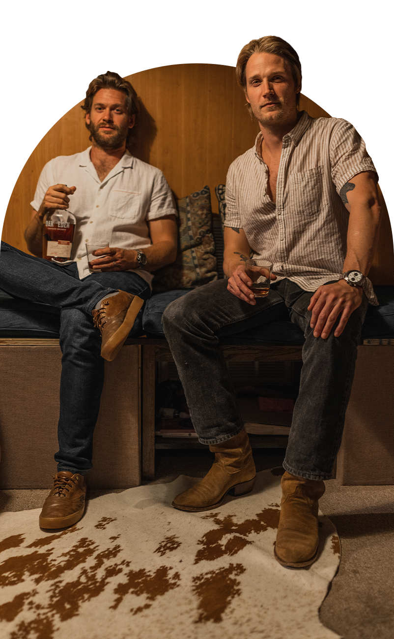 Jonathan Clay and Zach Chance of Jamestown Revival sitting against a wooden wall with cow skin rug at their feet, both holding a whiskey glass. The man on the left is holding a bottle of Oak & Eden’s barrel proof rye whiskey called Round Prairie Rye.