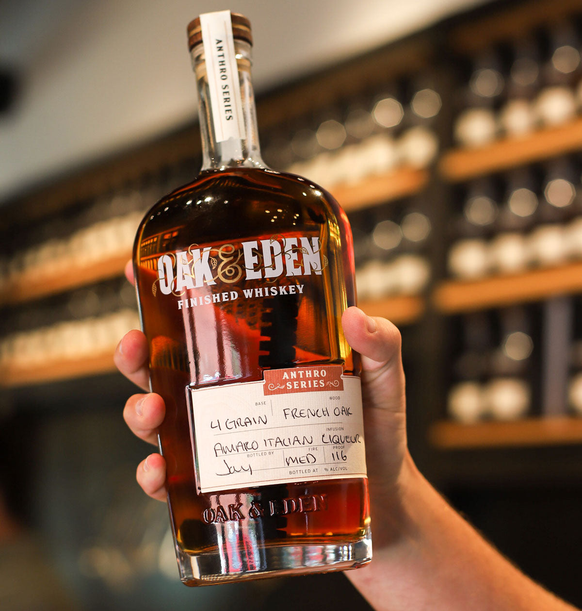 A hand is holding a bottle of Oak & Eden whiskey in front of a bar that has shelving lining the walls in the background.