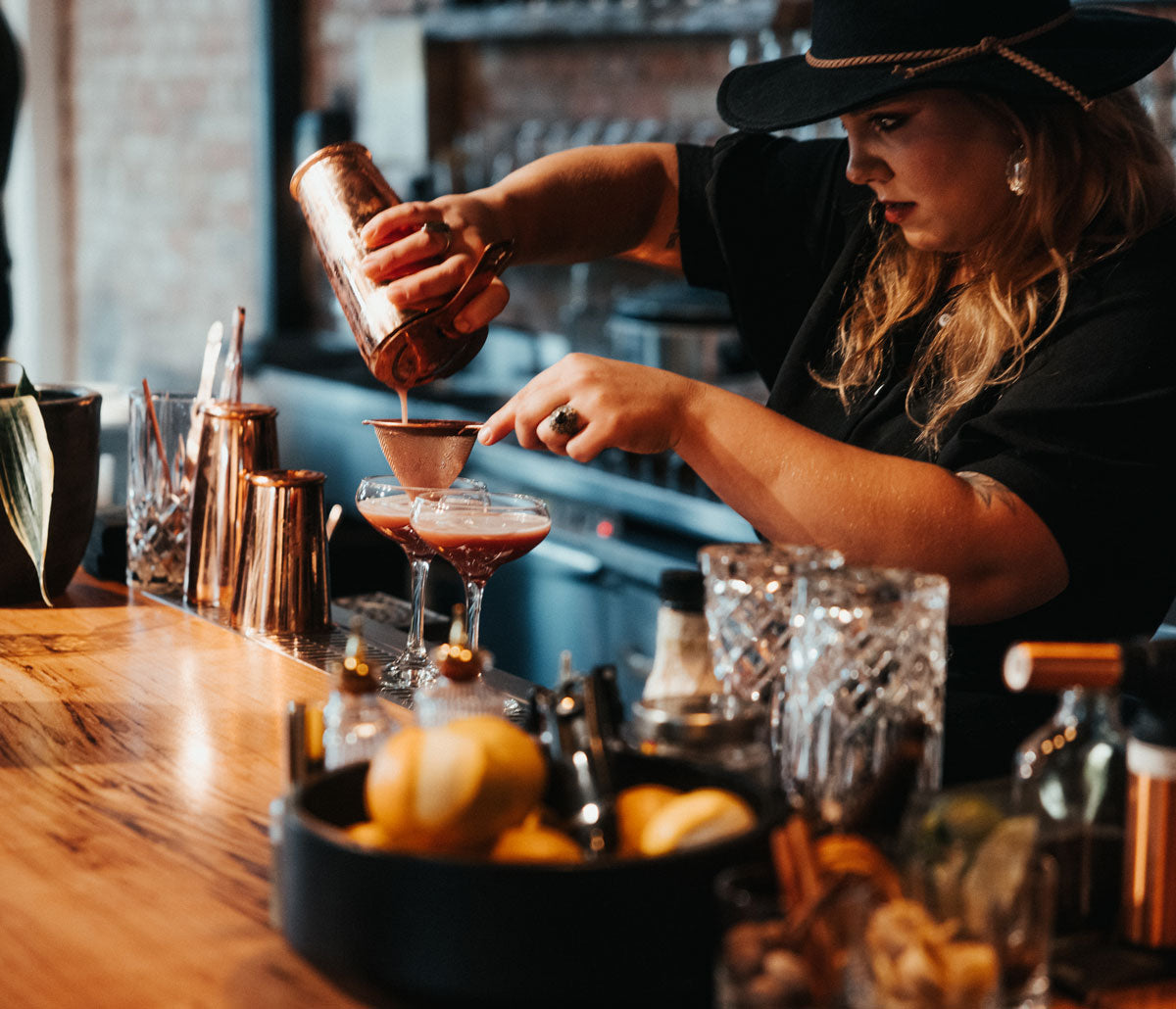 A women bartender is behind the bar, where she has just mixed a drink and is straining it into a cocktail glass. There is additional glass ware and other bar equipment lined up in front of her.